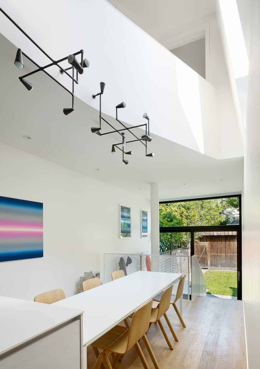 This informal dining area is in the middle of the kitchen and the staircase that has a white metal mesh railing that matches with the white table that is paired with wooden chairs and topped with an industrial style chandelier above it.