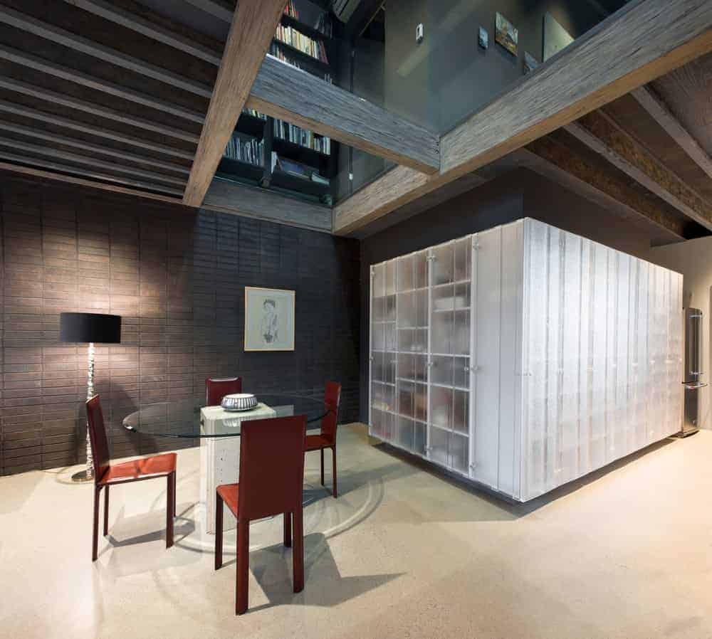 The round glass-top dining table is paired with red modern chairs that contrast the light hue of the industrial-style flooring that complements the stone brick walls and wooden ceiling with exposed wooden beams and glass panels.