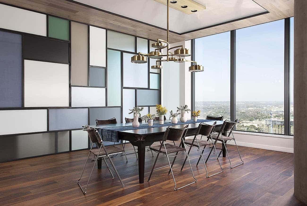 The brightness of this industrial-style dining room comes from the large glass wall that offers an amazing over-looking view. This brings in an abundance of natural lighting to the dark hardwood flooring contrasted by the colorful wall of panels and the silver legs of the chairs.