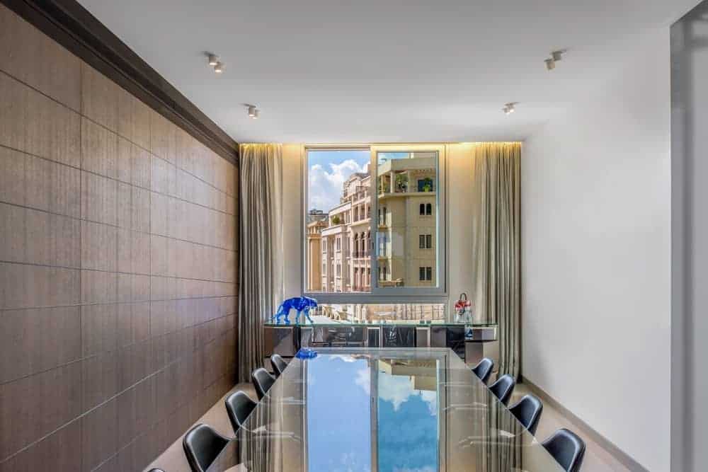 The industrial-style textured stone wall on the left provides an earthy tone contrast to the bright white wall on the other side as well as the white ceiling that is brightened by the window as well as the glass dining table.