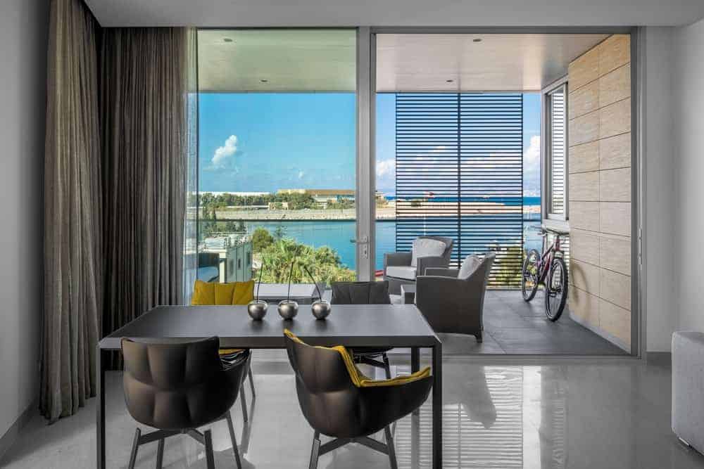 This simple dining area has a sleek industrial-style flooring that complements the dark gray modern dining table that is paired with black modern chairs that stand out against the light gray walls illuminated by the large glass sliding doors.