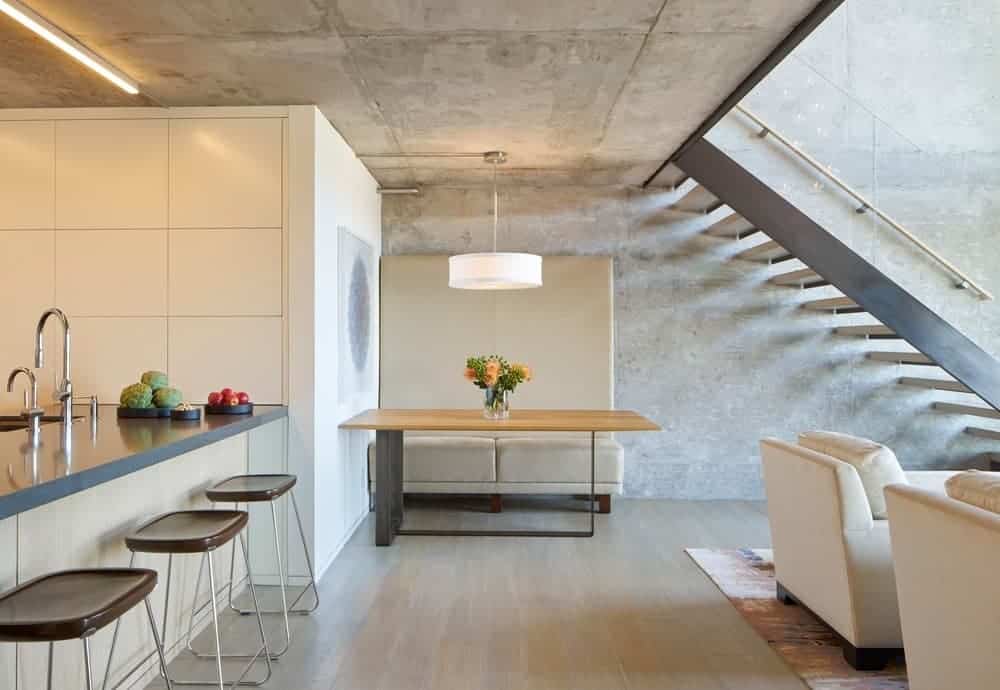 The simple informal dining area is tucked away in a corner by the kitchen and the living room. It has a beige cushioned bench with a large backrest that complements the gray concrete walls and ceiling that supports a white pendant light over the wooden table.