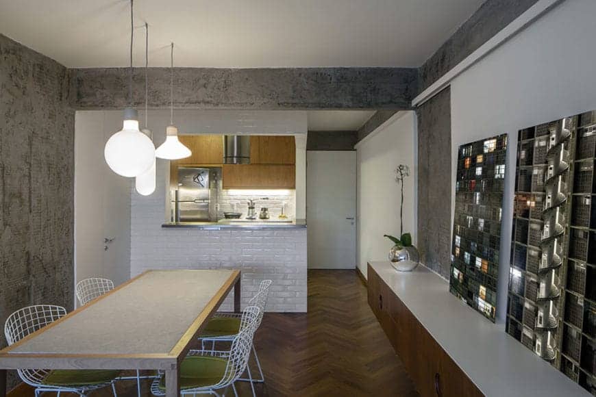The gray wooden dining table is paired with white metal outdoor-style chairs that stand out against the the dark hardwood flooring with a herring bone pattern as well as with the gray concrete walls adorned with photos.