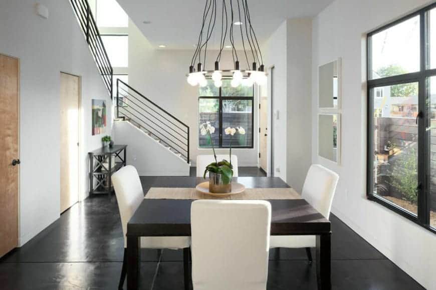 The white cushions of the dining chairs surrounding the black square dining table stands out against the black industrial-style flooring that is illuminated by the tall window and augmented by the industrial-style chandelier.