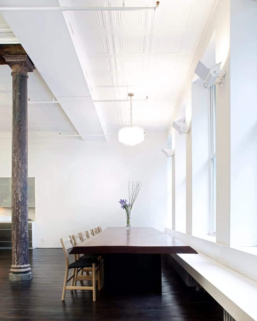 The large wooden dining table is paired with a built-in long bench on one side and a row of wooden chairs on the other side, These are all over a dark hardwood flooring and a high ceiling with exposed pipes.