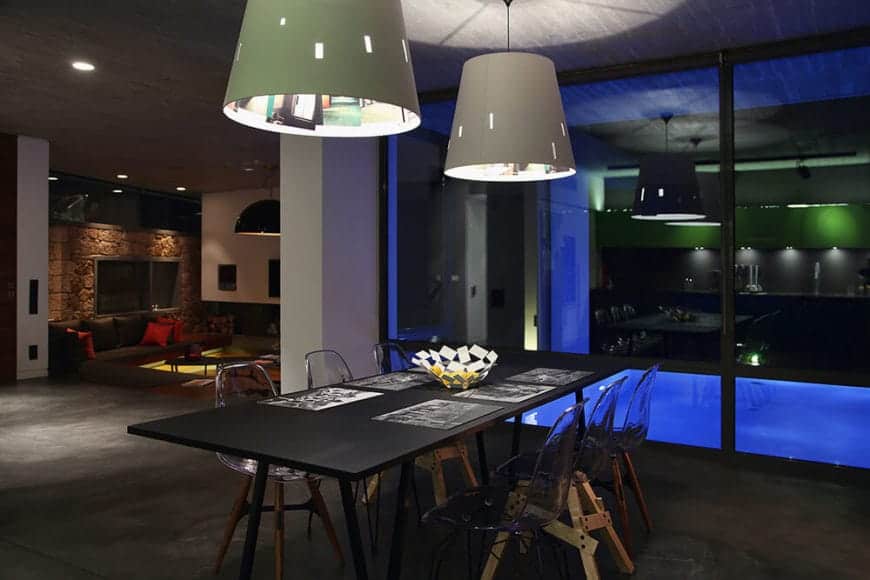 The bluish glow comes from the pool right outside the large glass wall by the head of the black wooden dining table that is surrounded by modern see-through chairs with wooden legs that stand out against the gray concrete flooring.