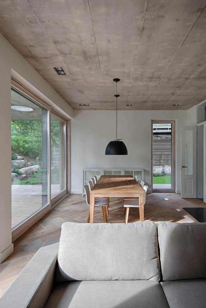 The simple wooden dining table stands out against the gray concrete ceiling that hangs a large dome pendant light with a black hue that contrasts the white modern dining chairs over a distressed hardwood flooring by the living room.