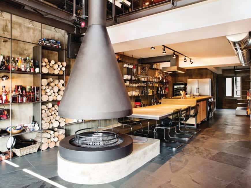 This dining area is in the middle of a large circular fireplace and the kitchen island all on a gray industrial flooring and a white ceiling that has exposed stainless steel ducts and vents. The dining area has a wooden table paired with modern chairs.