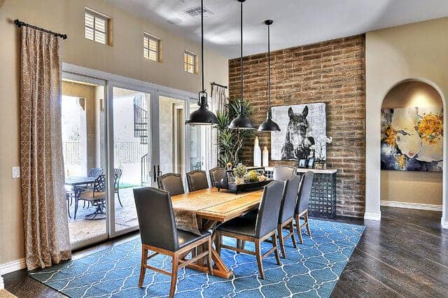 This dining room has an adobe brick wall with a dark gray hue that matches the hardwood flooring covered by a green patterned area rug underneath the wooden dining table paired with gray leather cushioned chairs.