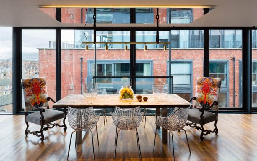 The simple wooden table matches the hardwood flooring that is illuminated by the large glass wall reflected by the mirror panel on the ceiling that support a row of pendant lights over the variety of chairs matched with the table.