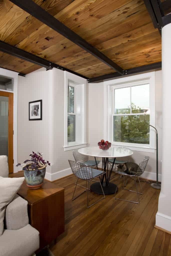 This small industrial-style dining area is in a nook alcove made by white walls with white framed windows and a wooden flooring and ceiling that has exposed metal beams with a contrasting black color matching the legs of the white dining table.