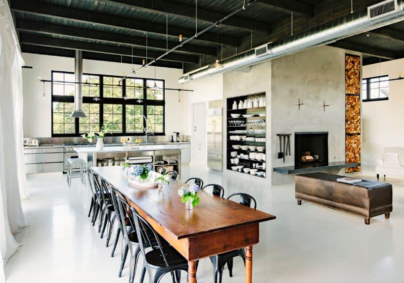 This large and airy dining area by the kitchen and living room has a long wooden rectangular dining table paired with dark metal chairs that stand out against the light hue of the industrial-style flooring paired with a black industrial-style ceiling.