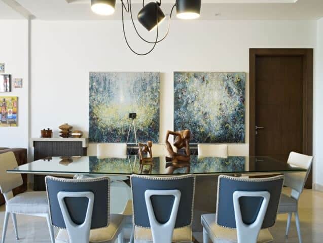 This dining room has a glass top dining table paired with modern chairs that have white cushions. These setup is topped with several pendant lights with exposed wiring that stands out against the white ceiling.