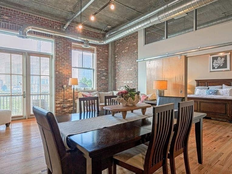 The elegant wooden dining set stands out against the industrial-style elements of this dining area like the red brick walls and the exposed pipes, vents and ducts of the gray high concrete ceiling above.