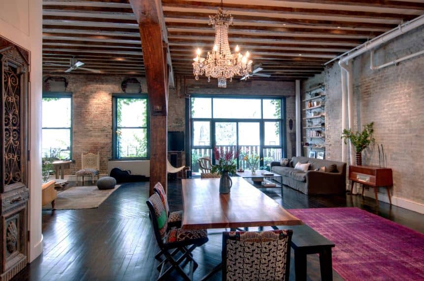 This dining area has a wooden ceiling with exposed wooden beams that match with the large column beam that supports it. This pairs well with the wooden table that is paired with a wooden bench near a purple area rug.