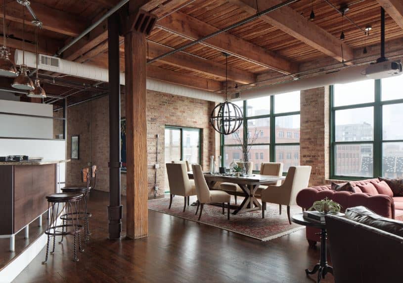 The brightness of this industrial-style dining room comes from the two large glass windows that dominate the red brick walls bringing in natural lights to the dark wooden dining table and its beige cushioned chairs topped with a spherical pendant light.