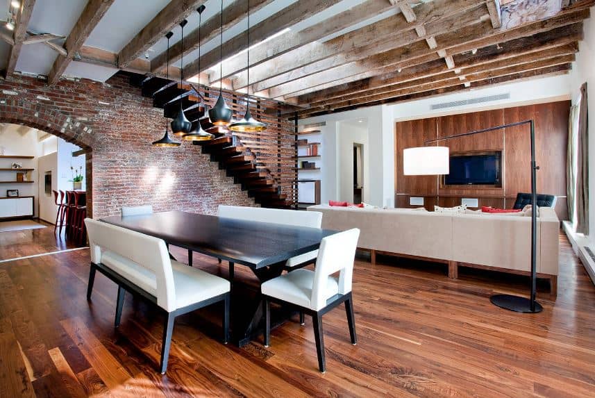 The redwood flooring is a complement to the red brick walls of this dining area behind the living room area. It has a contrasting black wooden table and white cushioned benches and chairs topped with a variety of pendant lights.