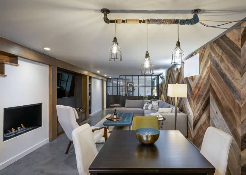 This small dining area beside the living room has the same wooden wall with a herring bone pattern and gray concrete flooring that contrasts the white cushions of the dining chairs of the dark wooden table topped with pendant lights.