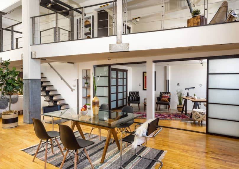 The dining table has a glass top and wood frame paired with modern chairs over a patterned area rug. This setup is given an industrial-style background of concrete columns, metal beams and metal railings as well as a hardwood flooring.