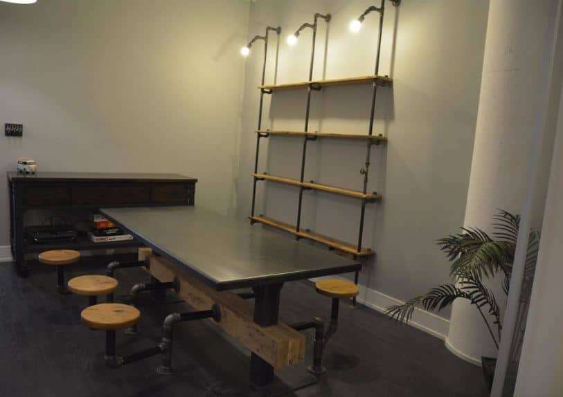 This industrial-style dining room is dominated by a black dining table with dark iron legs supporting a wooden beam that is the main attachment of the stools surrounding the table that has water pipes for legs. This is mirrored by the shelving on the light gray wall.
