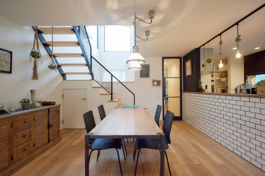 The simplicity of this dining room gives it warmth and a homey quality to its hardwood flooring that pairs with the wooden dining table topped with industrial-style pendant lights and a low wall with a brick pattern.