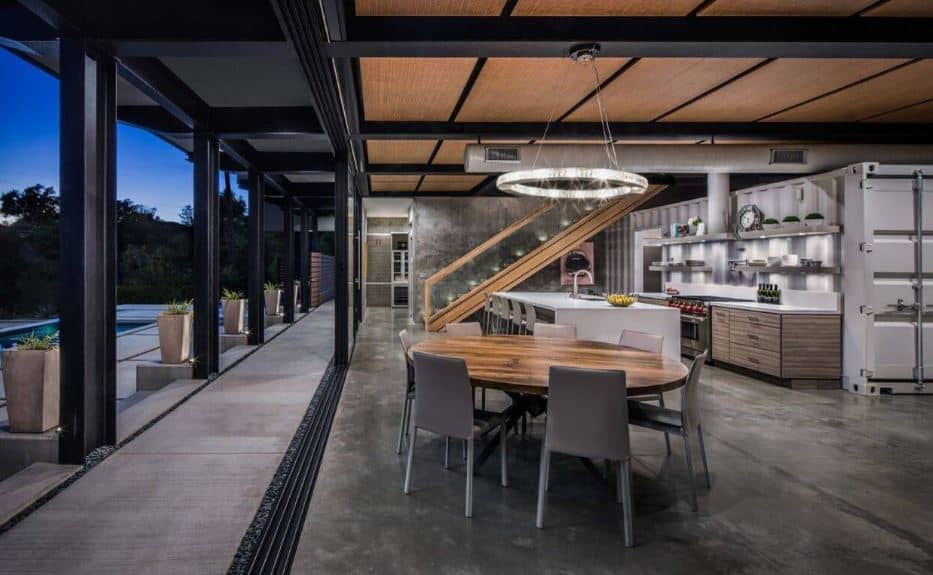 The circular wooden table is paired with light gray modern dining chairs that are matched with the gray industrial flooring topped with a wooden ceiling with exposed metal beams and a background of a repurposed shipping container at the kitchen.
