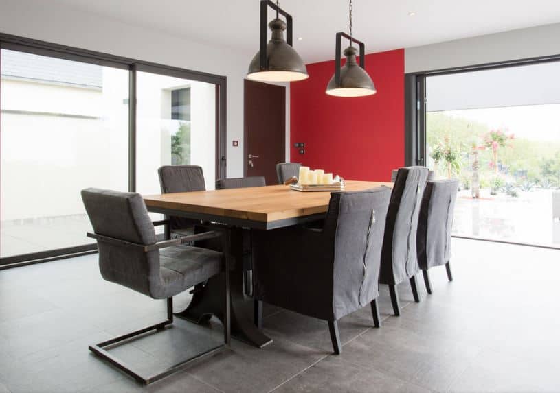 The dining table has dark metallic legs that match the dark gray velvet cushions of the chairs. The table has a thick slab of wood on top that stands out against the gray industrial-style flooring and the dark pendant lights above.