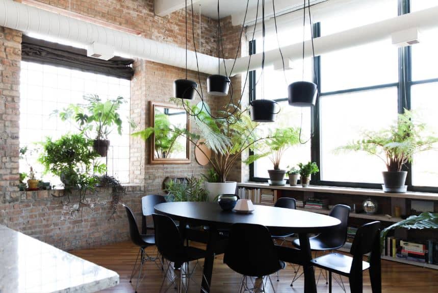 The oblong black dining table is paired with equally dark modern dining chairs. This setup is complemented by the dark hardwood flooring and the red brick walls brightened by large windows and white air ducts.