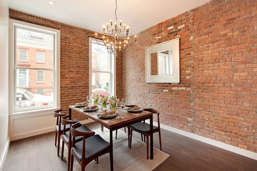 This dining room has a brick wall that complements the dark hardwood flooring matching with the wooden dining set. This is contrasted by a white ceiling that matches with the white frames of the windows that bring in natural lights.