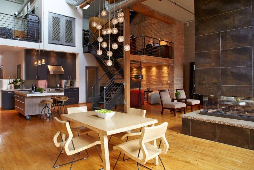 The light wooden dining table is matched with dining chairs of the same hue that stands out against the hardwood flooring with a slightly darker hue. This is complemented by a high ceiling with exposed ducts and vents as well as pendant lights.