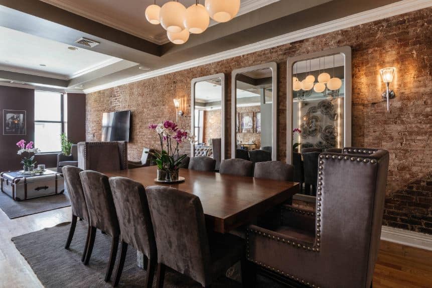 The red brick walls of this industrial-style dining room is adorned with a trio of wall-mounted mirrors with gray frames that complement the dark brown cushions of the dining chairs and the dark wooden dining table.
