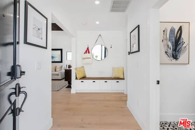 As you enter this bright Farmhouse-style foyer, you will see a a sitting area for the waiting guests. It has a built-in bench with a wooden top embedded into the white wall. This bench has storage areas at the bottom for the shoes and hooks on its wall for the coats, hats and bags.