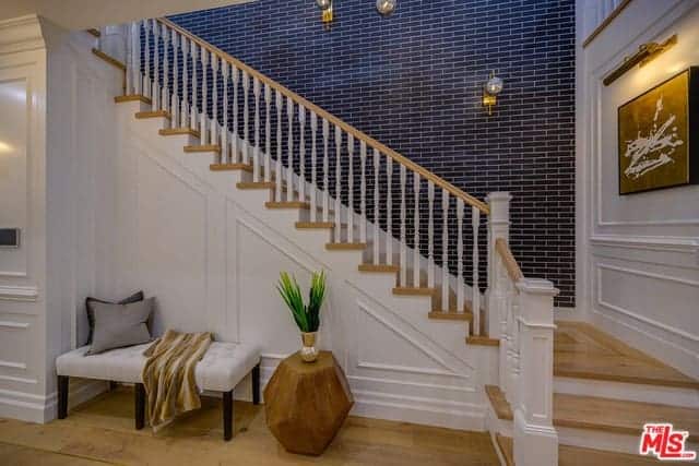 This foyer has a lovely view of the wooden staircase that has white railings and wooden steps contrasted by the black wall with a brick wall finish. Below the stairs is a comfortable waiting area with a cushioned bench that blends with the white wooden walls.