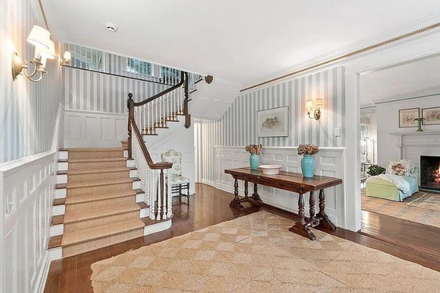 The dark hardwood flooring of this elegant foyer is mostly covered by a brown woven area rug that matches with the carpeting of the stairs. These are augmented by the white wainscoting contrasted by the dark wooden console table bearing flower vases.