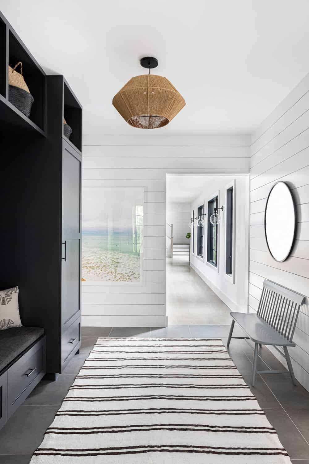The white ceiling of this Farmhouse-style foyer is adorned with a pendant light that has a woven basket hood that provides a rustic element to the dark wooden mudroom on the left side with a cushioned seat. Opposite this is a light gray wooden bench against the white plank wall.