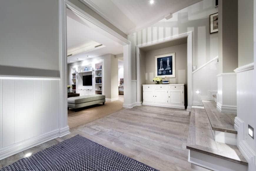 The simple patterned area rug marks the entrance to this simple Farmhouse-style foyer that has a hardwood flooring matching the steps of the stairs. This is complemented by the white walls and white shed ceiling with bright recessed lights.