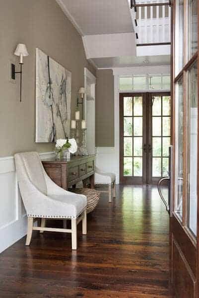 The dark wooden main that has glass panels on it matches with the dark hardwood flooring of the Farmhouse-style foyer that has a comfortable cushioned chair on the side for the waiting guests.
