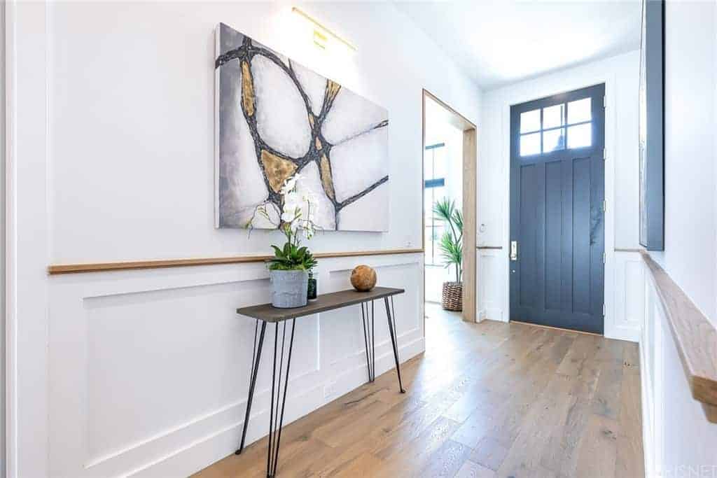 This bright white foyer is decorated with a simple wood-top console table with thin iron legs. It stands out against the white wooden finish of the wall that is accented with a colorful painting above. These are all illuminated by the transom windows of the door.