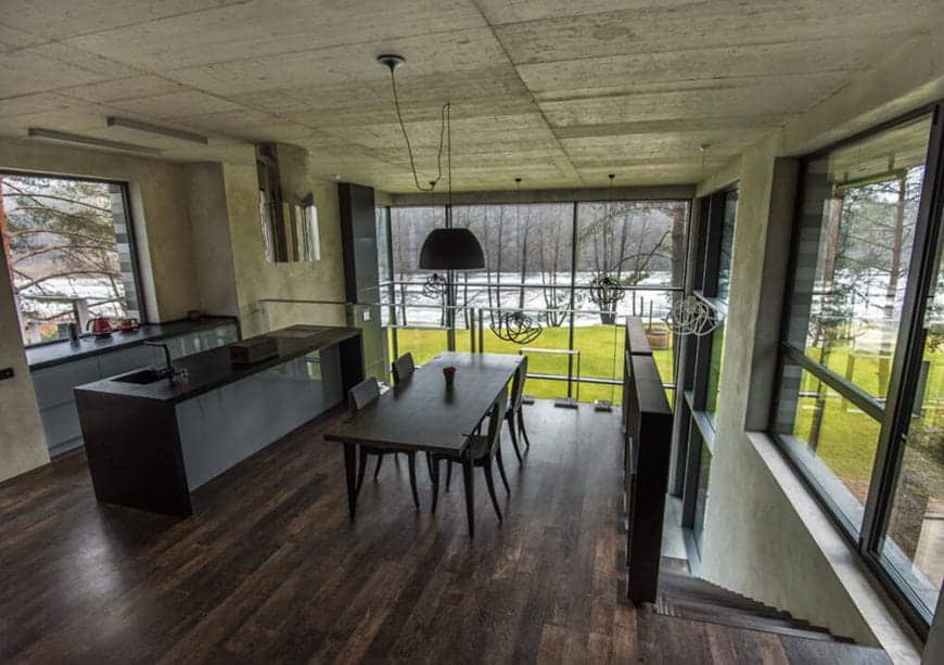 This is a second floor kitchen with a hardwood flooring that blends with the kitchen island and an industrial-style gray concrete ceiling and walls brightened by the large glass windows that feature a wonderful landscape scenery outside.