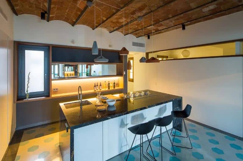 This industrial-style kitchen has a unique ceiling made of brick with a small arches to it. This is matched with an equally unique flooring that has gray and green hexagonal tiles arranged in a polka dot pattern. This is a nice background for the simple waterfall kitchen island and small peninsula.