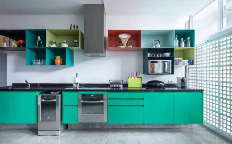 This cheerful kitchen has a green floating modern peninsula with a black countertop. Above it are wall-mounted open shelves with different colors that stand out against the white walls and the appliances that match with the gray concrete flooring.
