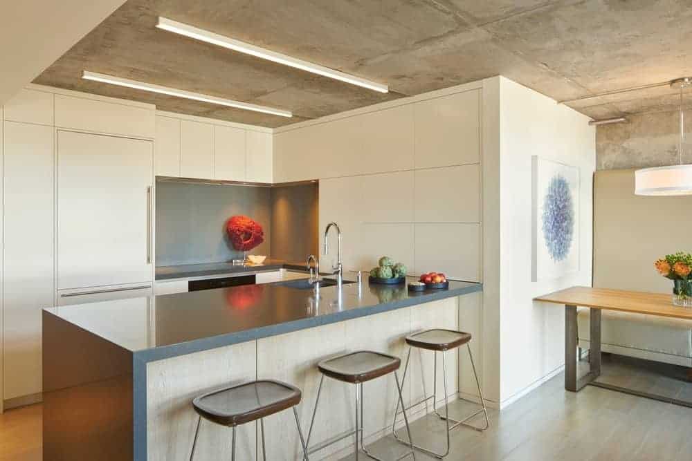 The ceiling is made of gray concrete that matches with the waterfall kitchen island. The ceiling has a couple of beige lights that match the kitchen cabinets of the L-shaped peninsula that houses the various appliances.