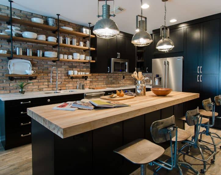 The industrial-style pendant lights are a great complement for the butcher block countertop of the black kitchen island matching with the peninsula that houses the stainless steel appliances complemented by the red brick walls.