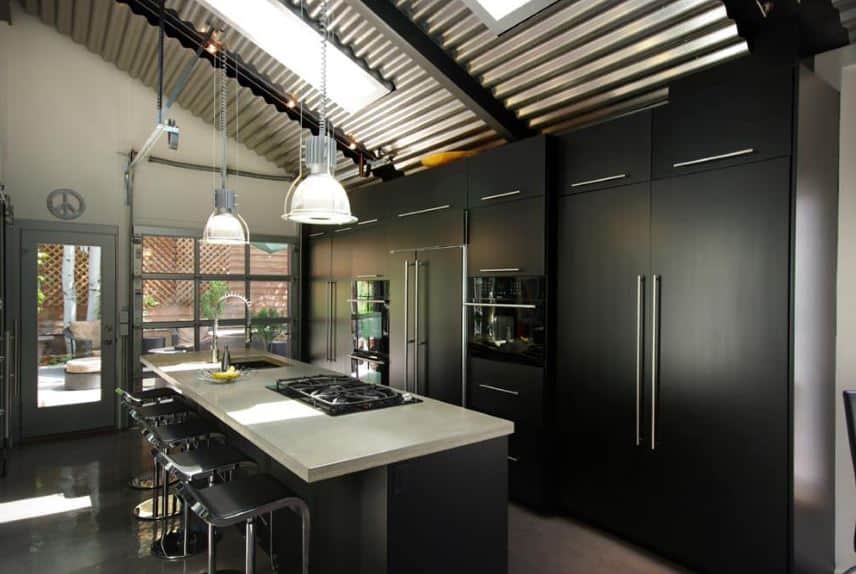 The shed ceiling is made of stainless tin roofing sheets that are held together by exposed dark beams. This ceiling also has sky lights that cast natural lights down onto the modern kitchen that is mostly black and gray.
