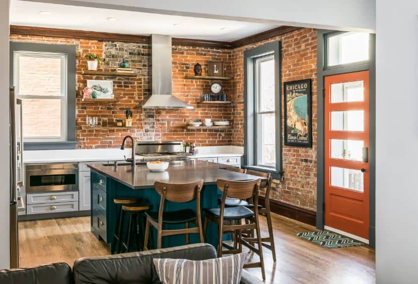 The green cabinets and drawers of the kitchen island stands out against the red brick walls of the kitchen peninsula that has gray shaker cabinets and drawers housing the stainless steel appliances and its vent hood above.