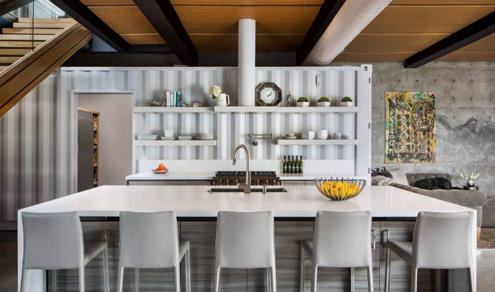 The white countertop of the peninsula goes well with the white wall that looks like a part of a shipping container. This is paired with white shelved and a white vent hood and air duct on the wooden ceiling with exposed black beams.