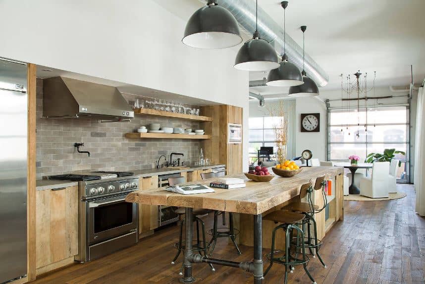 The large wooden kitchen island has a thick butcher block countertop that is supported by legs made of water pipes. This goes well with the exposed vent and duct of the white ceiling as well as the gray backsplash of the wooden peninsula.
