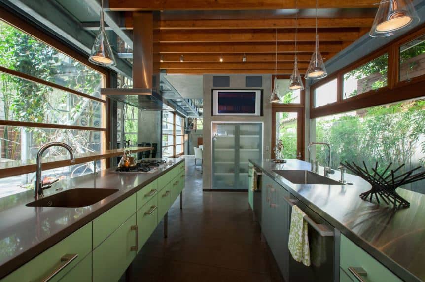 This narrow kitchen is flanked on both sides with glass walls that offer a nice green landscape scenery outside. This matches with the green modern cabinets of the two peninsulas with gray countertops that goes well with the wooden ceiling and its exposed wooden beams.