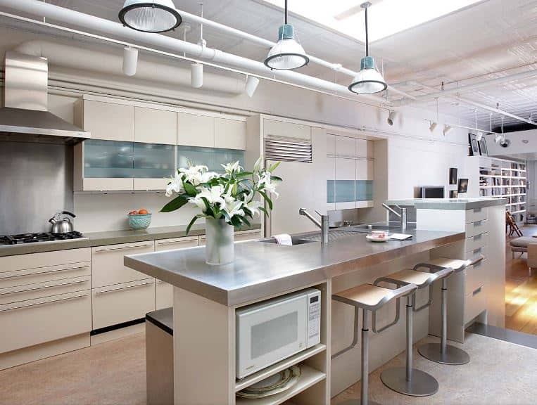 The brilliant white ceiling of this kitchen has exposed white pipes and tubes for the wiring of the spot lights and pendant lights hanging over the kitchen island that has a second level countertop to serve as a bar paired with modern stools.