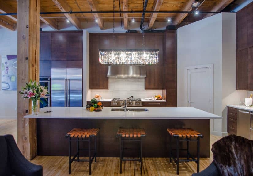 There is a large wooden column that connects the hardwood flooring and the wooden ceiling that has exposed wooden beams of the same hue as the flooring. These are a nice framing for the dark brown kitchen island and peninsula.